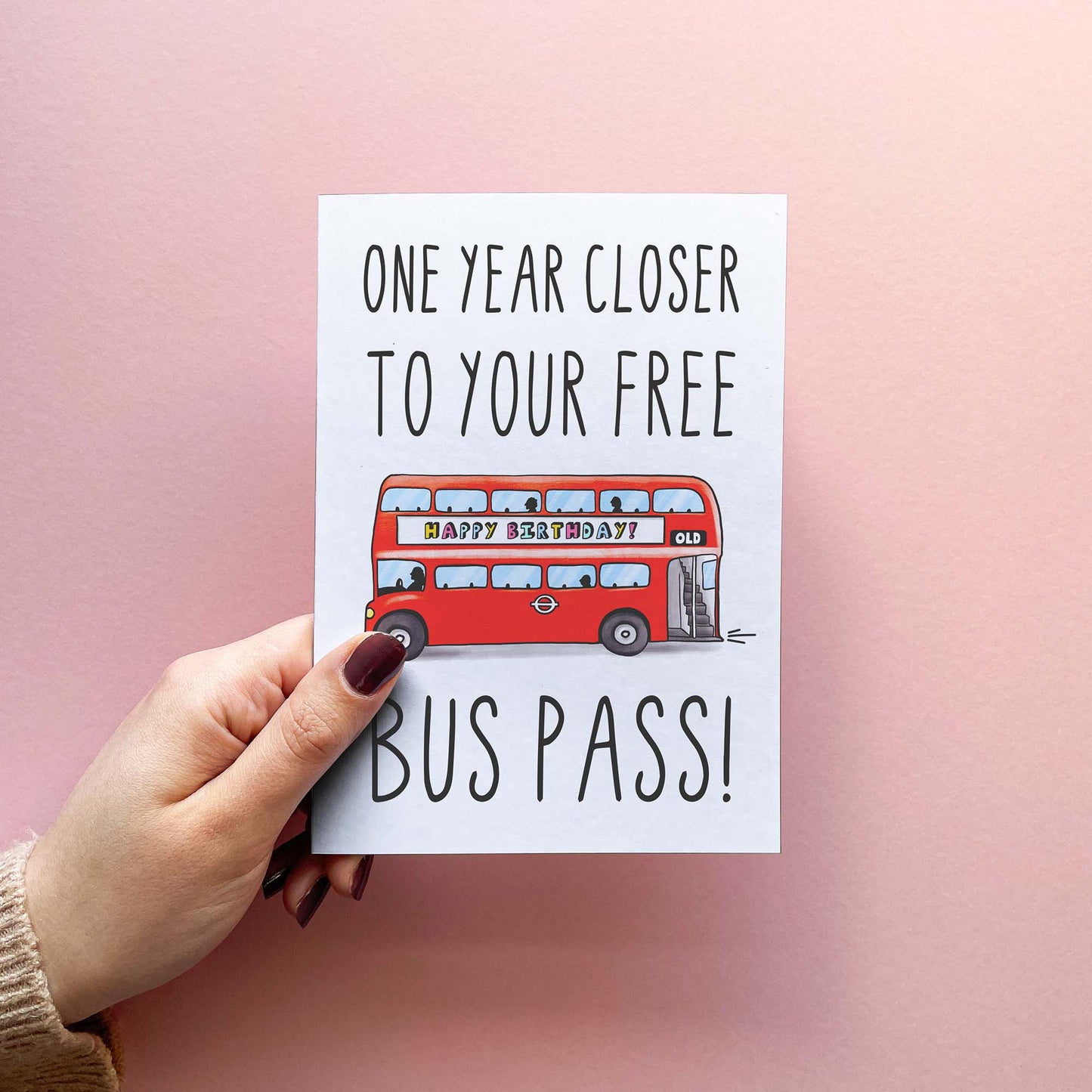 Cheeky birthday card reading 'one year closer to your free bus pass!' in handwritten font. Illustrated with red London bus displaying 'OLD' route number and 'happy birthday' advert. Includes Kraft envelope. Card is being held in hand for this photo.