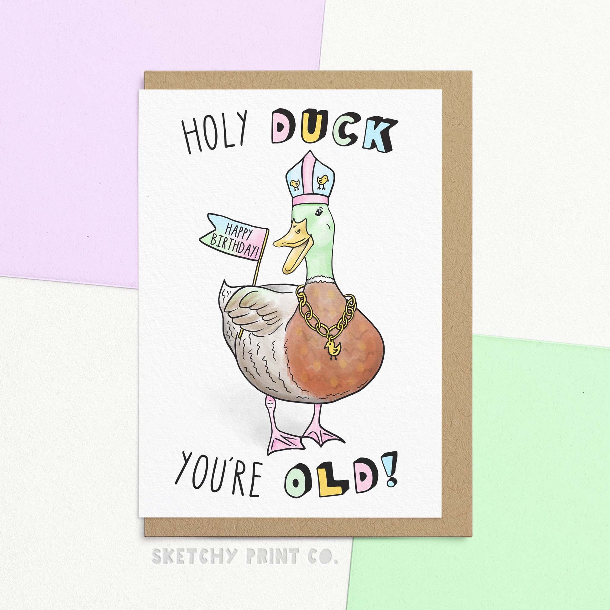 Funny birthday card featuring duck in mock-religious attire holding 'Happy Birthday' banner, text reads 'Holy Duck You're old!'. Shown with a Kraft Envelope.