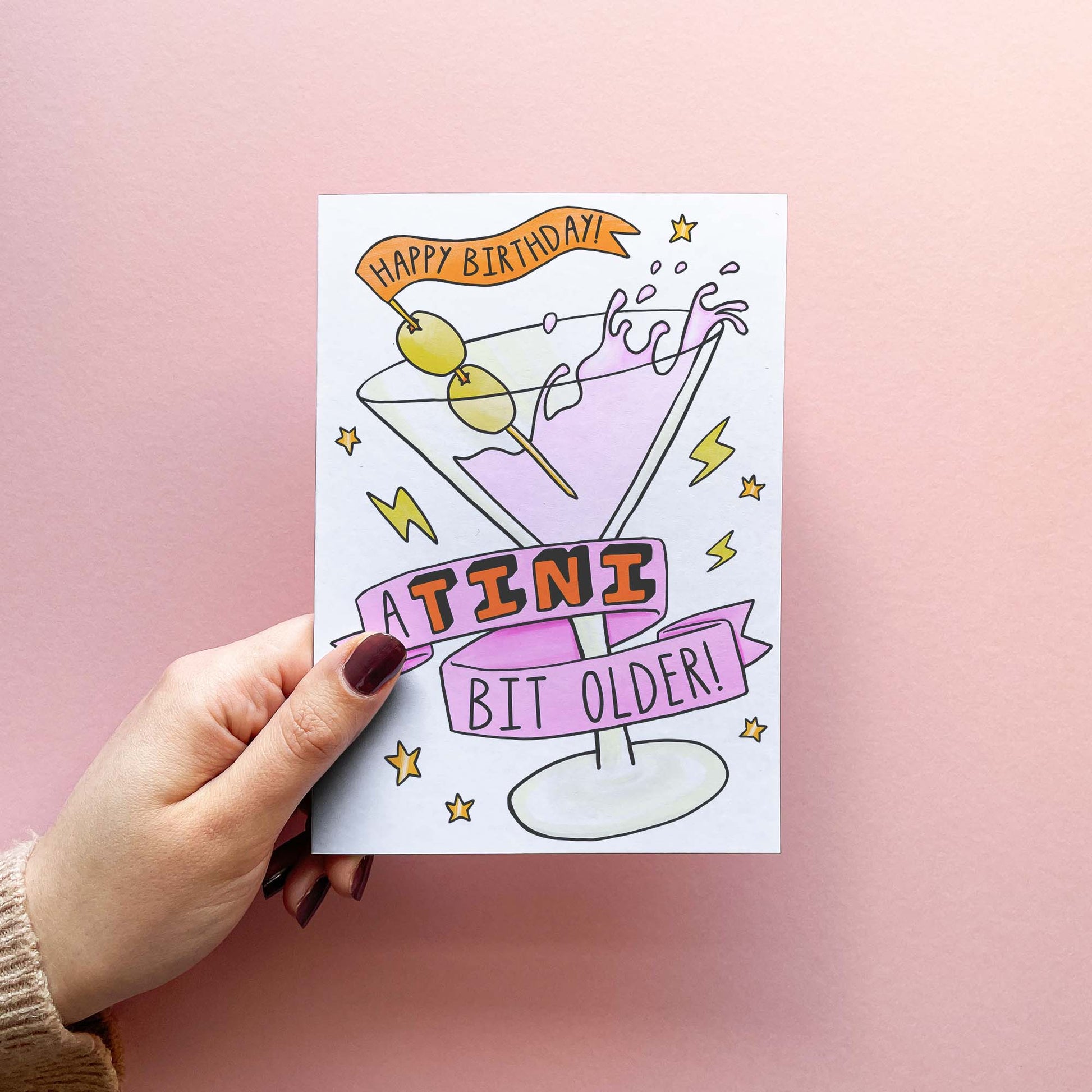 Stylish birthday card with martini illustration reading 'a tini bit older'. Cocktail stick says 'Happy Birthday!'. Shown in hand.