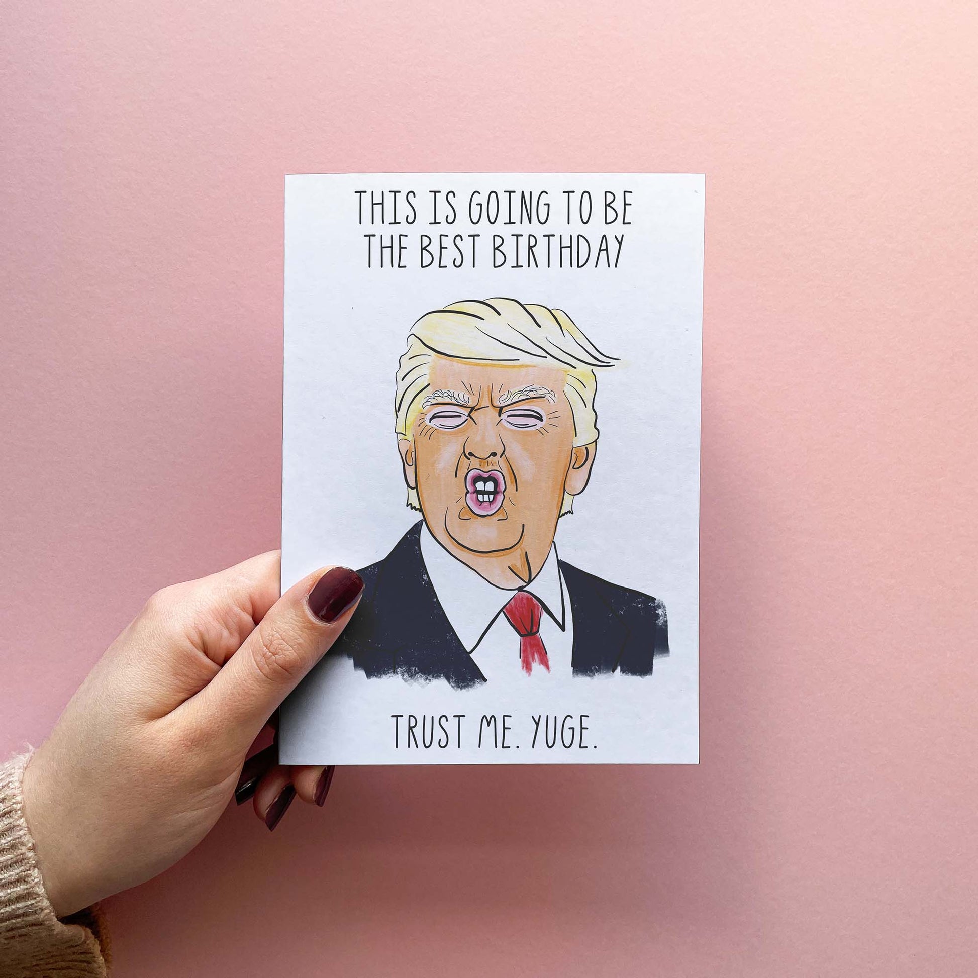 Funny birthday card featuring Trump illustration and reading 'this is going to be the best birthday. trust me. yuge.'. Shown In Hand.