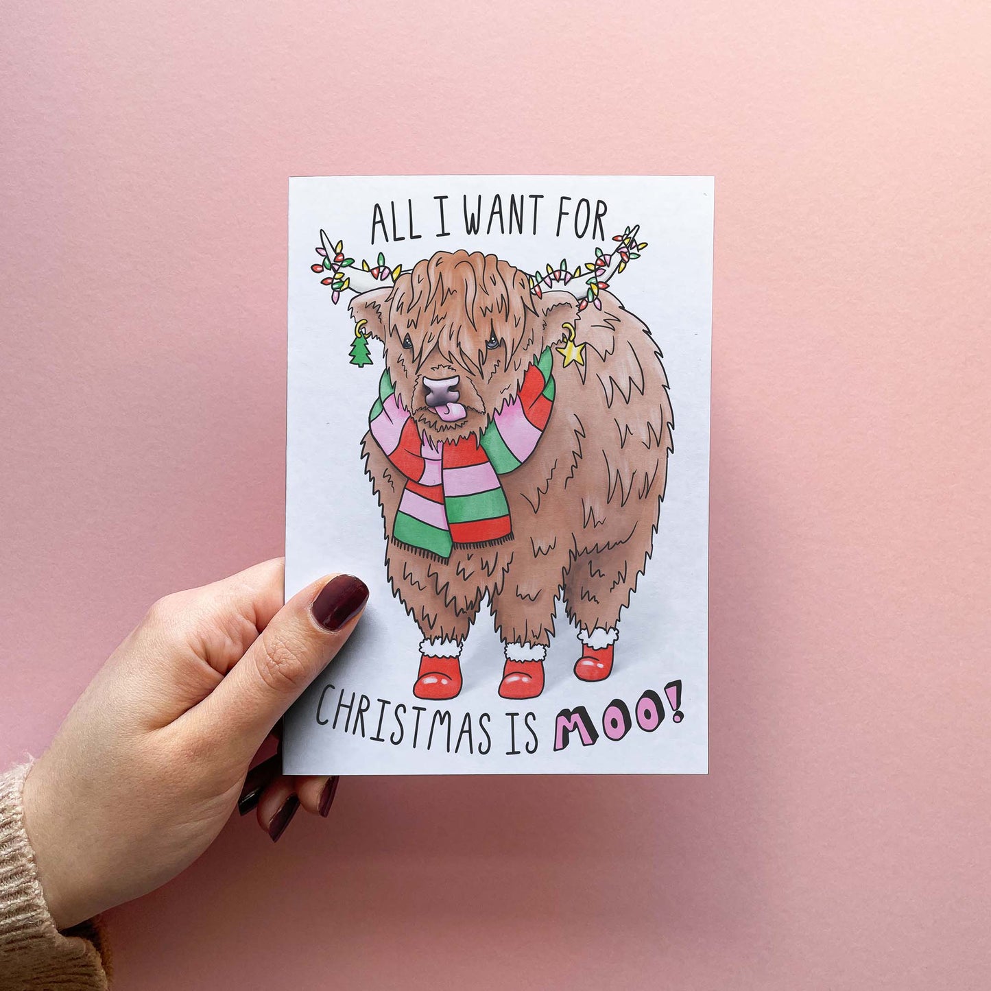 Funny Christmas card with cute Highland cow illustration and text pun 'all I want for Christmas is Moo!'. Shown in hand.