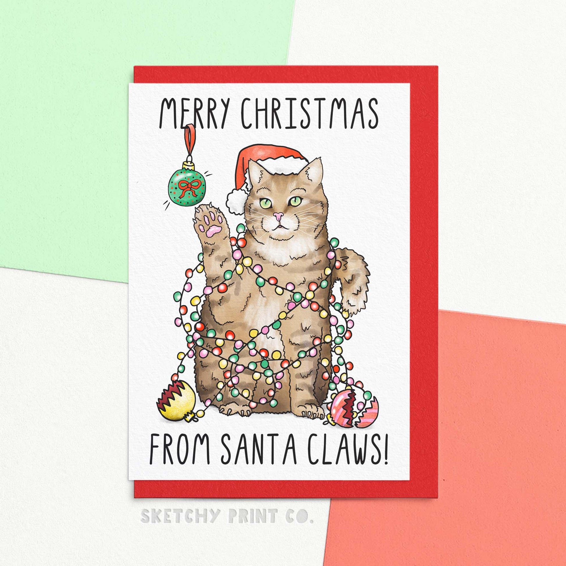 unny Christmas card showing cute cat tangled in Christmas lights with broken baubles, text reads 'Merry Christmas from Santa Claws!'. Shown with envelope.