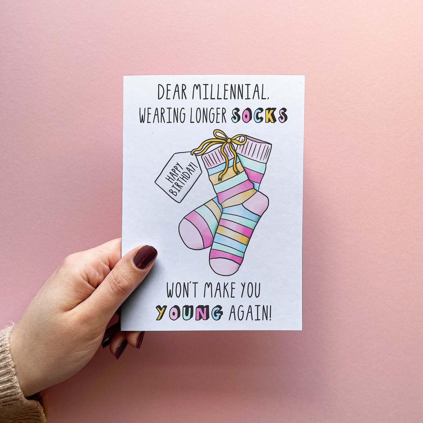 Funny birthday wishes for best friend or sibling. Card Reads Dear millennial, wearing longer socks won't make you young again! With an illustration of socks with a tag saying Happy Birthday! The card is held in hand in front of a pink background.