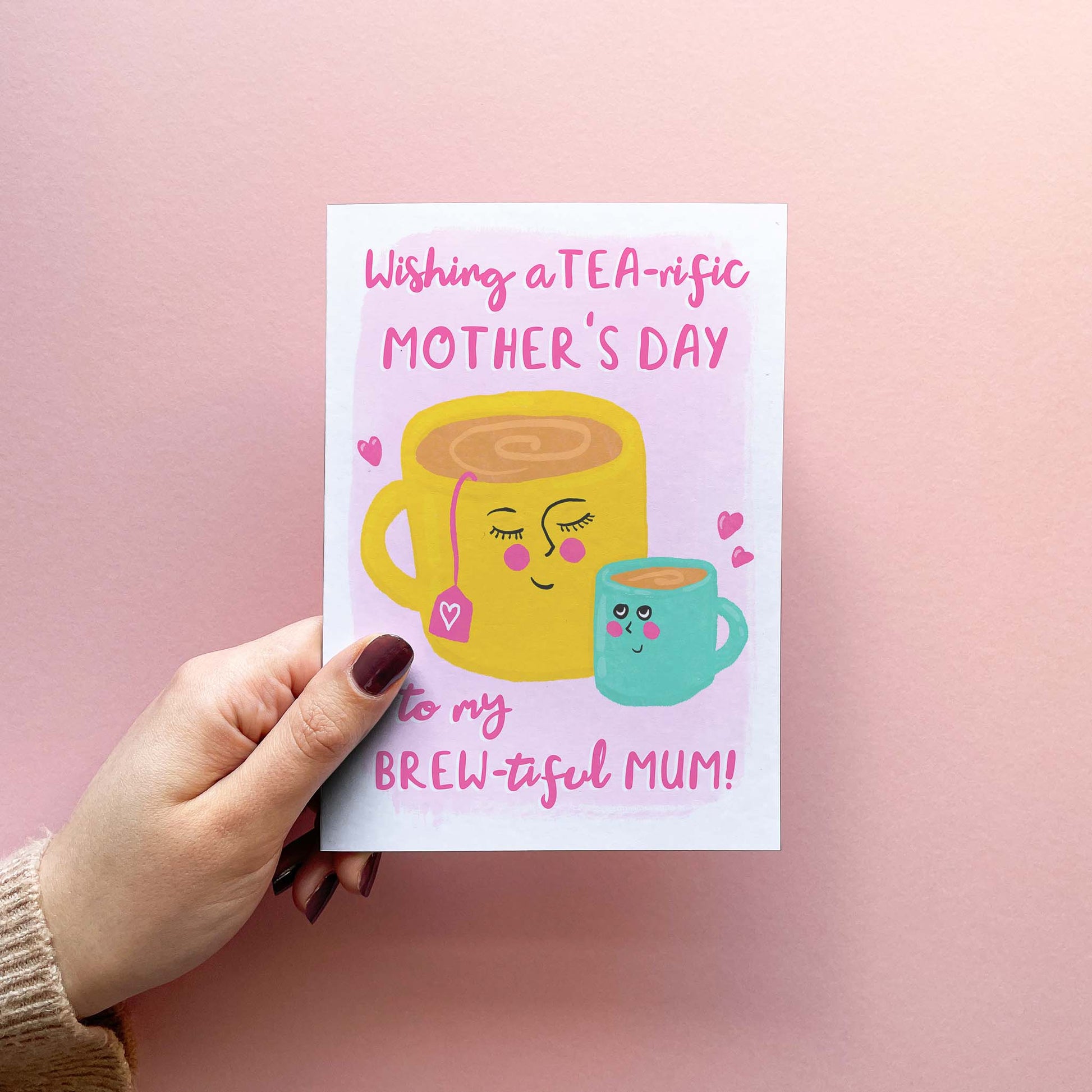 Cute and funny mother's day card for your tea loving mum featuring a modern colourful illustration of a mum and child cup of tea. With text reading "wishing a tea-riffic mother's day to my brew-tiful mum!". Card is shown being held.