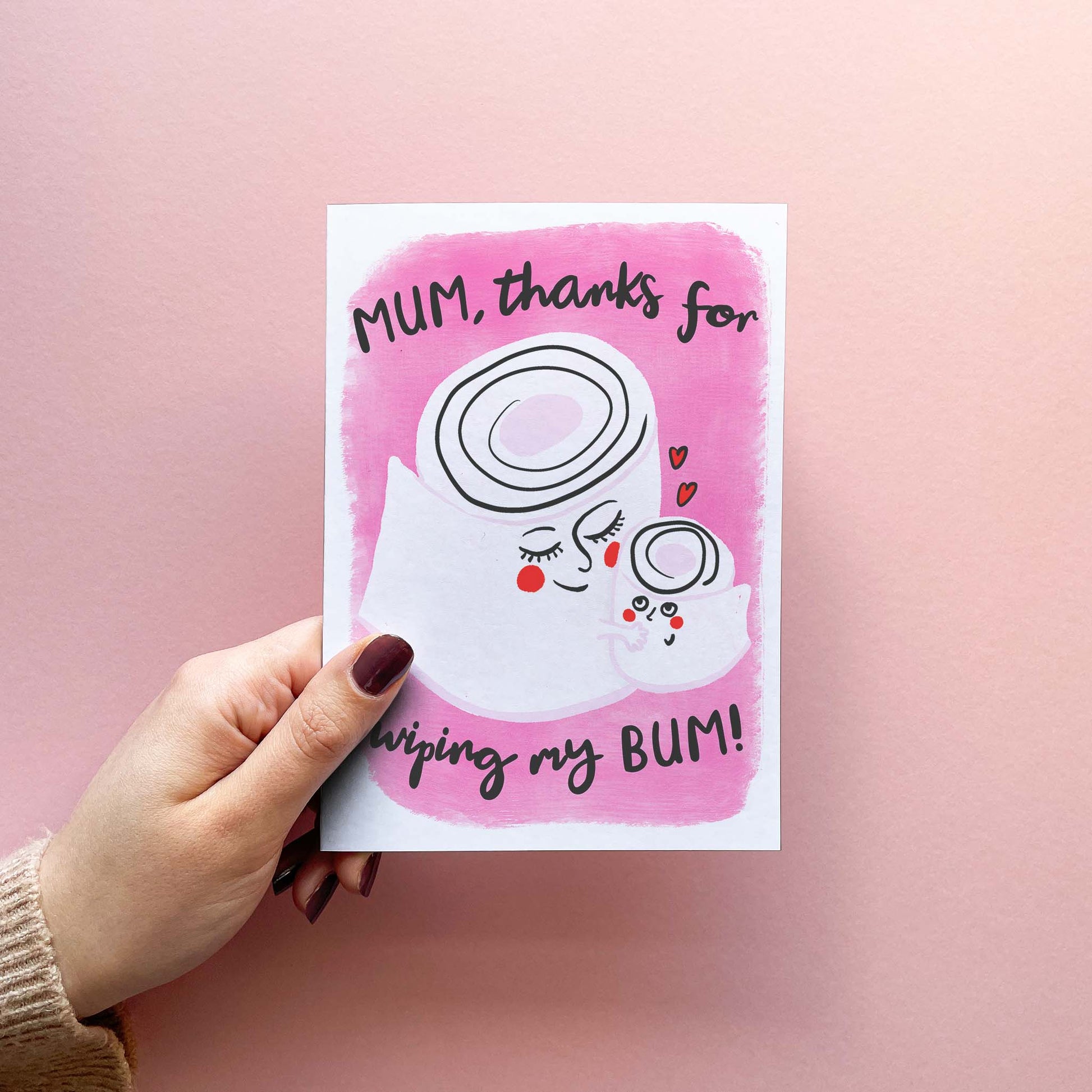Funny and cute Mothers Day card for Mum, featuring a modern whimsical illustration of mum and child cartoon toilet rolls on a pink background with text  reading "Mum, thanks for wiping my bum!". Card is shown being held.