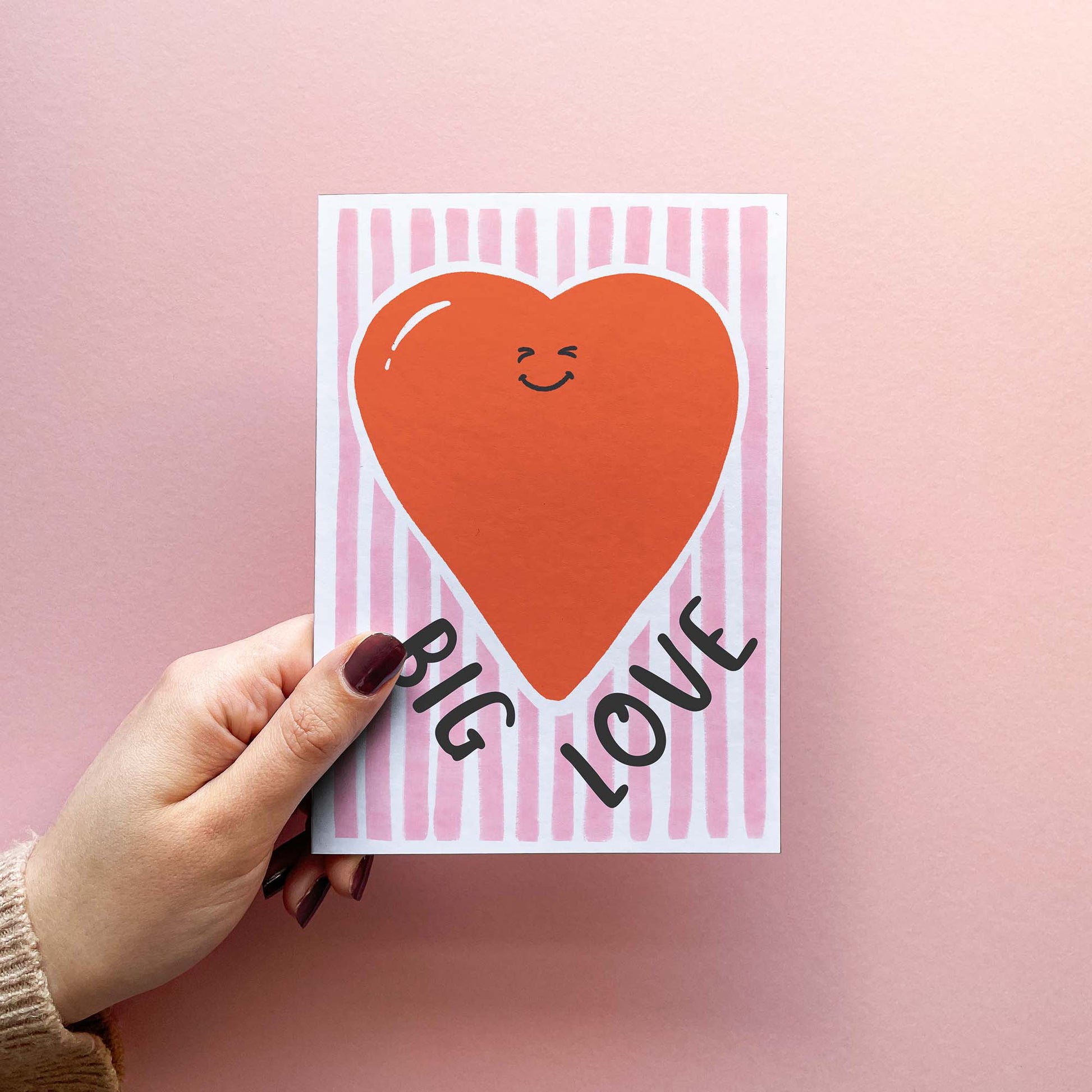 Hand holding a cute 'Big Love!' greeting card with fun, hand-lettered text and a smiling red heart on a stylish pink striped background.