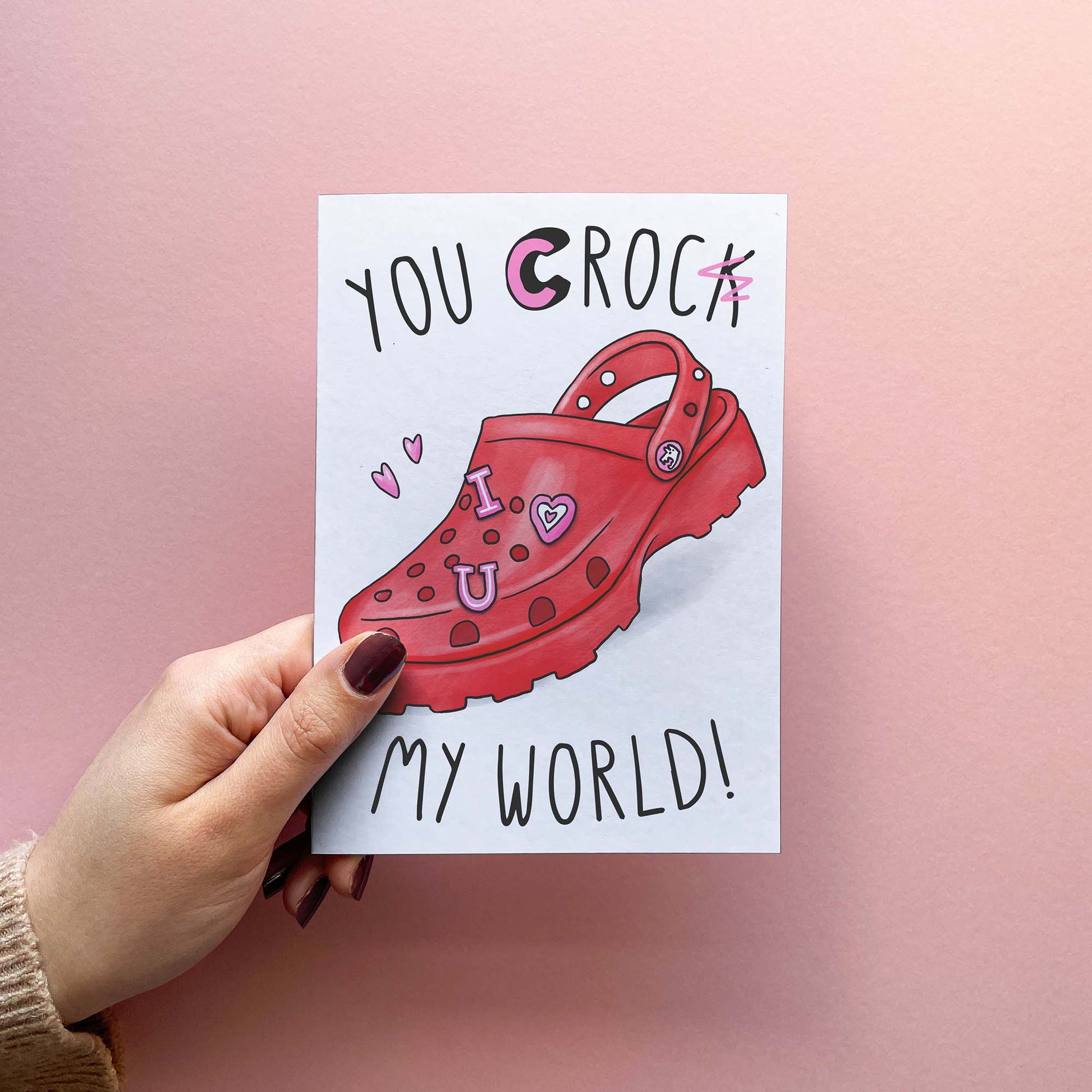 Funny anniversary card for husband or wife. Funny Valentine's Day card reading you croc my world! with the k scribbled out. Card is held in hand in front of a pink background.